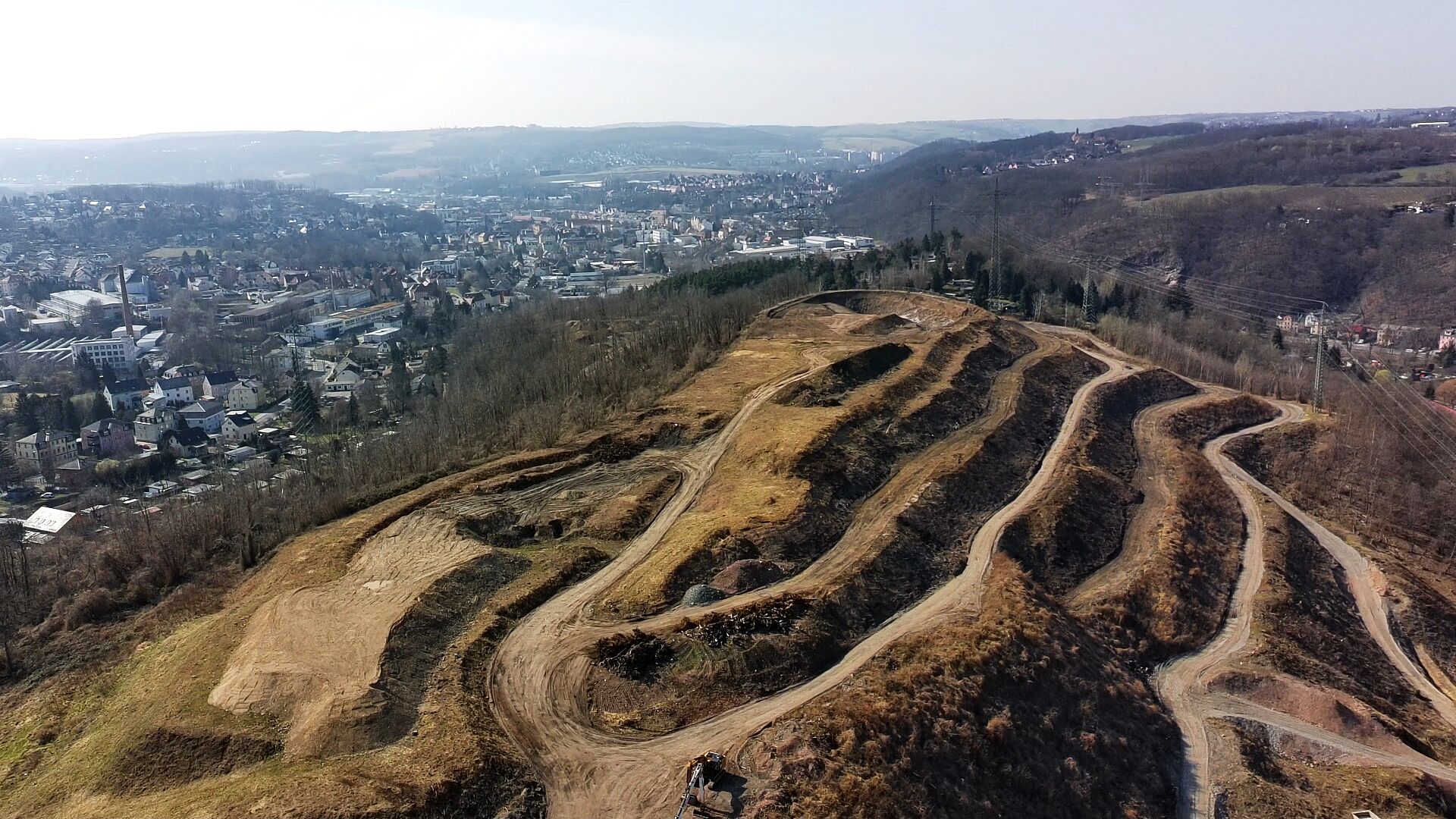 Die terrassierte Halde in ganzer Ausdehnung, dahinter der Ort und die Hügel des Tals.